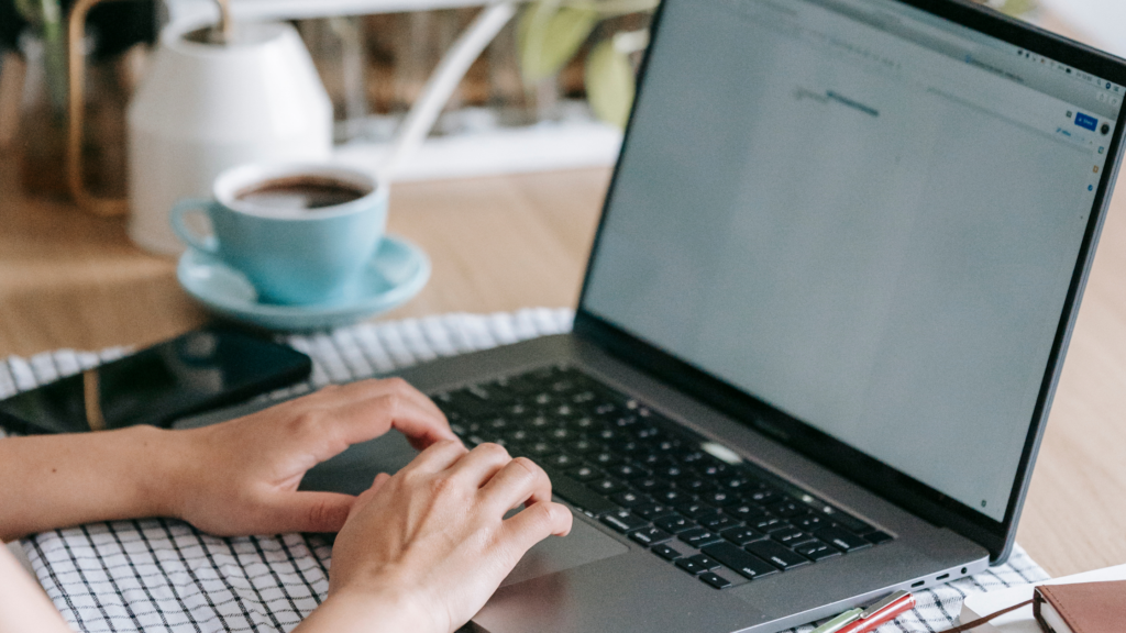 Looking over a woman's shoulder as she types on a generic laptop in Google Docs. Only her hands are visible hovering over the trackpad. She also has a coffee mug full in the background.